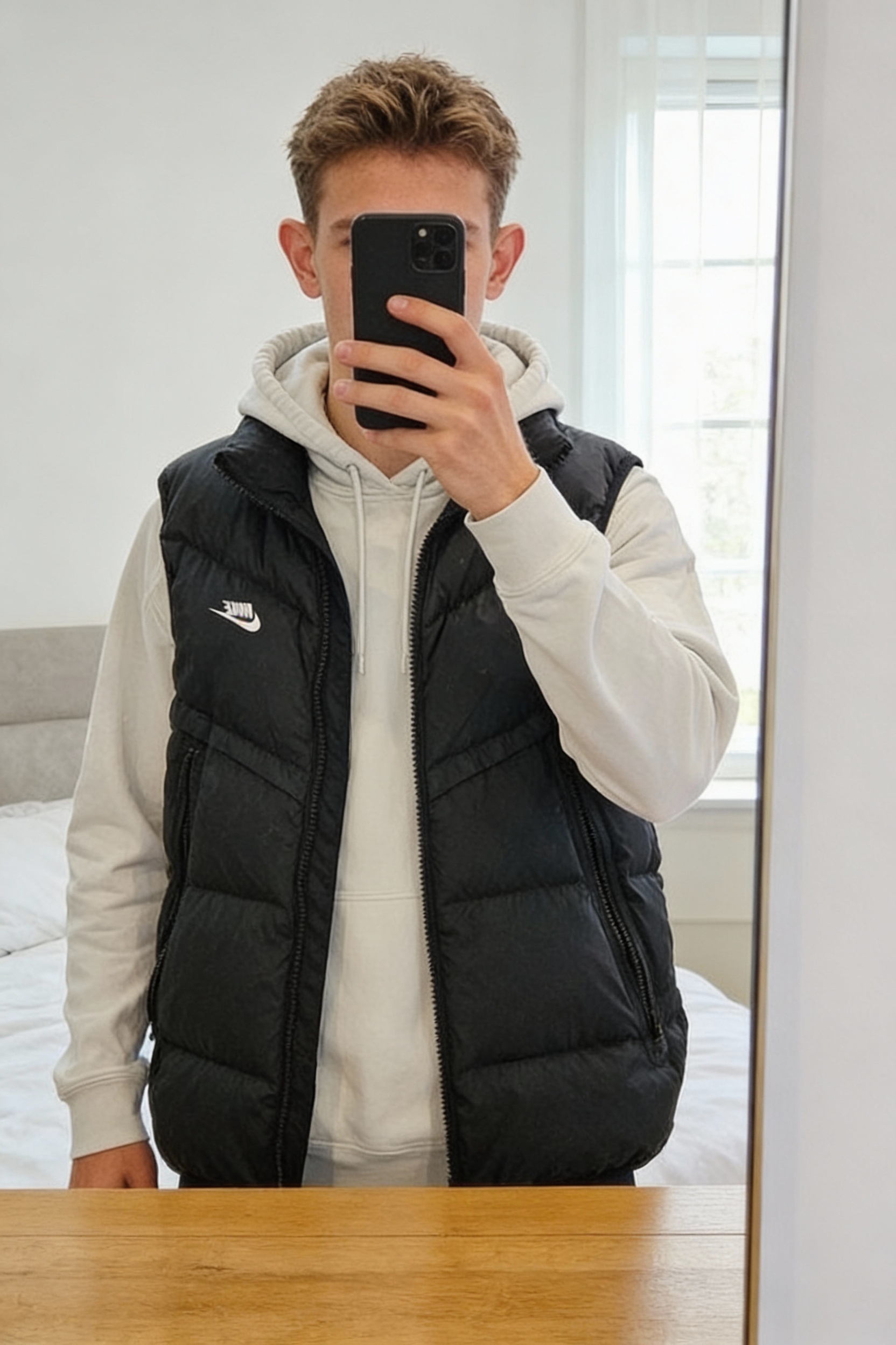 Sans manche Nike et sweat blanc debout, face à la caméra, dans une chambre à la lumière douce, avec une lumière naturelle filtrant par la fenêtre.