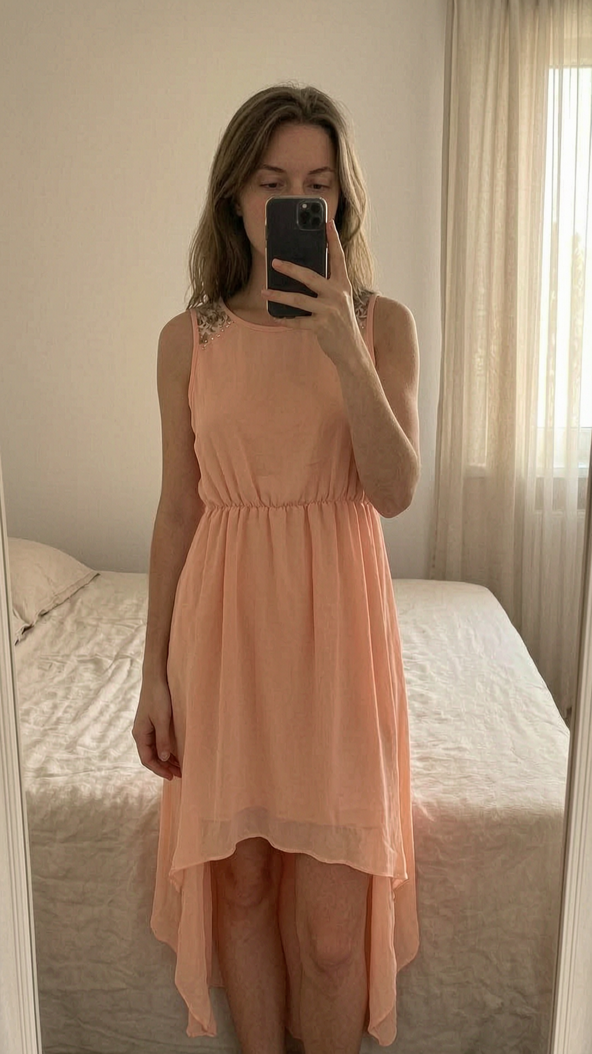 Robe saumon asymétrique debout, face à la caméra, dans une chambre à la lumière douce, avec une lumière naturelle filtrant par la fenêtre.