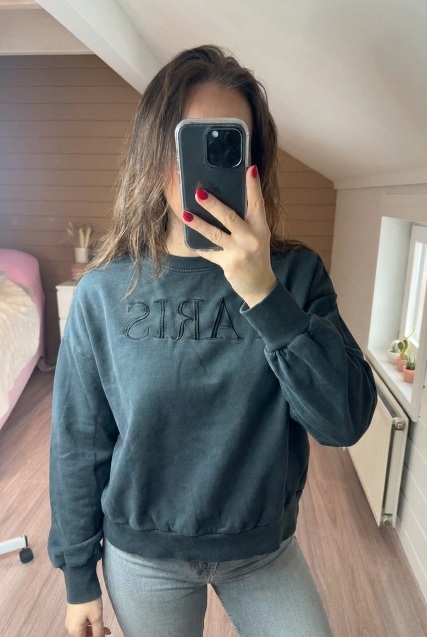 Pull only noir écriture Paris gris foncé large col rond court debout, face à la caméra, dans une chambre à la lumière douce, avec une lumière naturelle filtrant par la fenêtre.