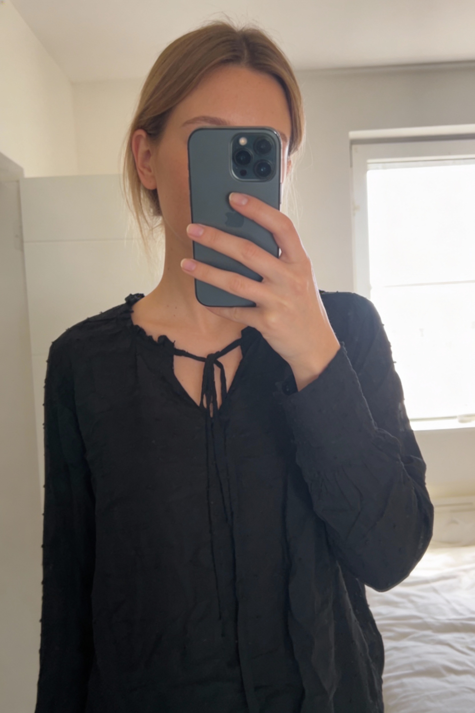 Blouse plumetis noir neuve S debout, face à la caméra, dans une chambre à la lumière douce, avec une lumière naturelle filtrant par la fenêtre.