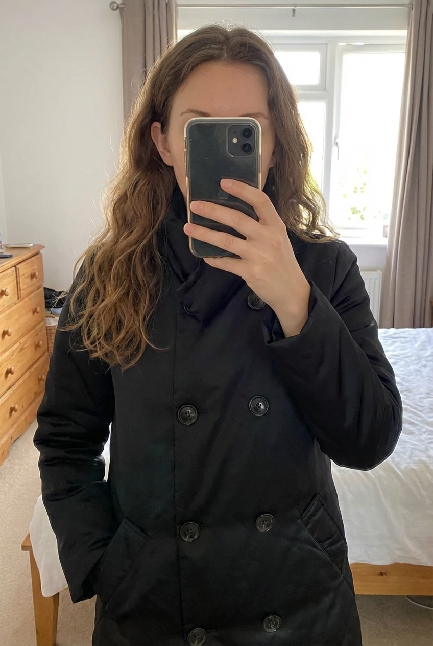 Manteau femme long matelassé noir Camaïeu femme – coupe droite, chaud, élégant taille L debout, face à la caméra, dans une chambre à la lumière douce, avec une lumière naturelle filtrant par la fenêtre.