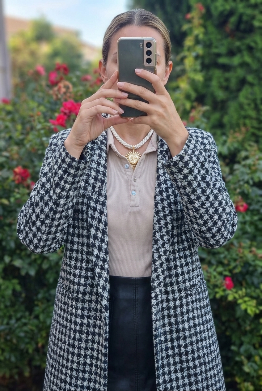 Une personne en Sublime manteau pied-de-poule noir et blanc – chic et intemporel cambrant légèrement le dos pour une pose dynamique, en extérieur, dans un cadre naturel entouré de verdure et de lumière douce.
