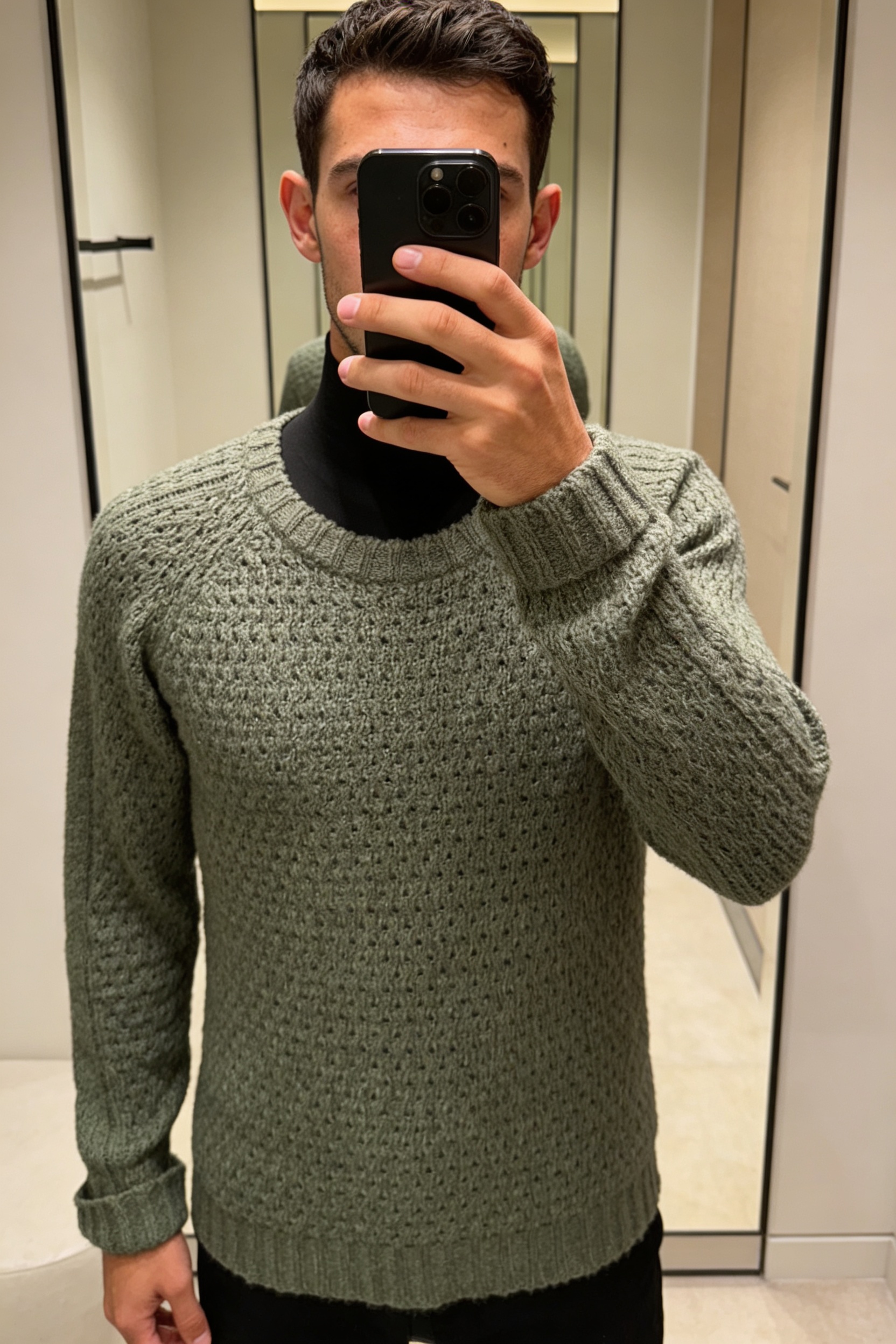 Una persona in Maglione Lavorato Verde Salvia - Taglia L Cod.3189 in piedi, rivolto verso la fotocamera, in un camerino in stile boutique con specchio e decorazioni neutre.