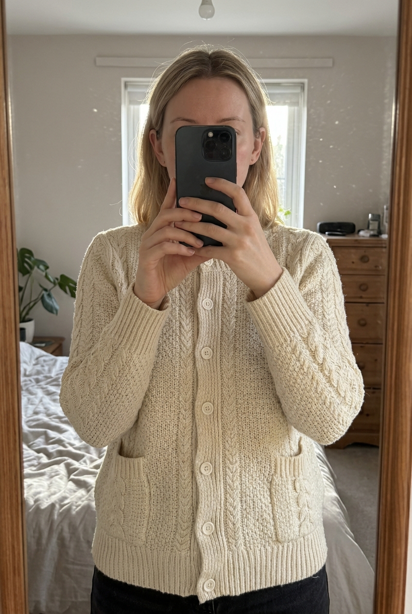 Une personne en Cardigan Torsadé Aran Sweater Blanc crème Aran Craft L debout, face à la caméra, dans une chambre à la lumière douce, avec une lumière naturelle filtrant par la fenêtre.
