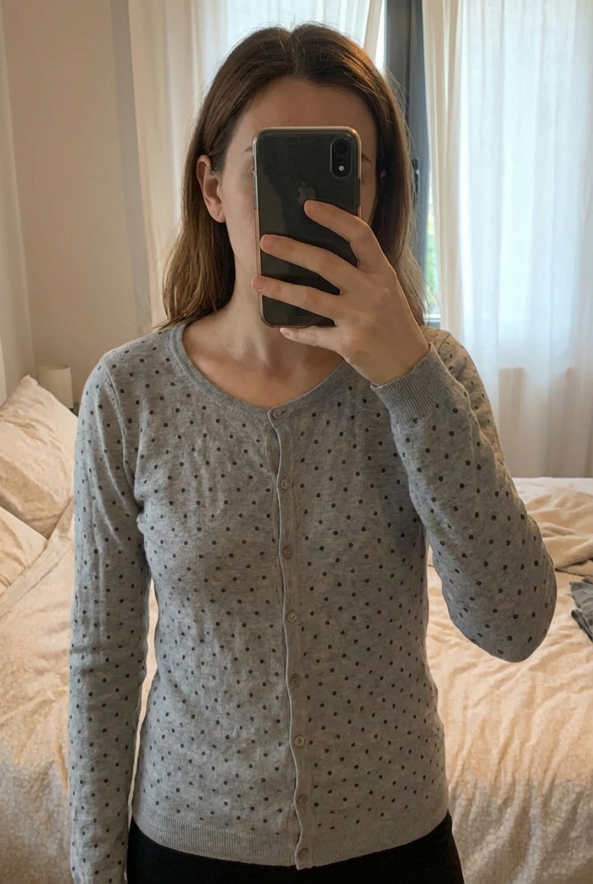 Gilet gris à pois noir femme debout, face à la caméra, dans une chambre à la lumière douce, avec une lumière naturelle filtrant par la fenêtre.