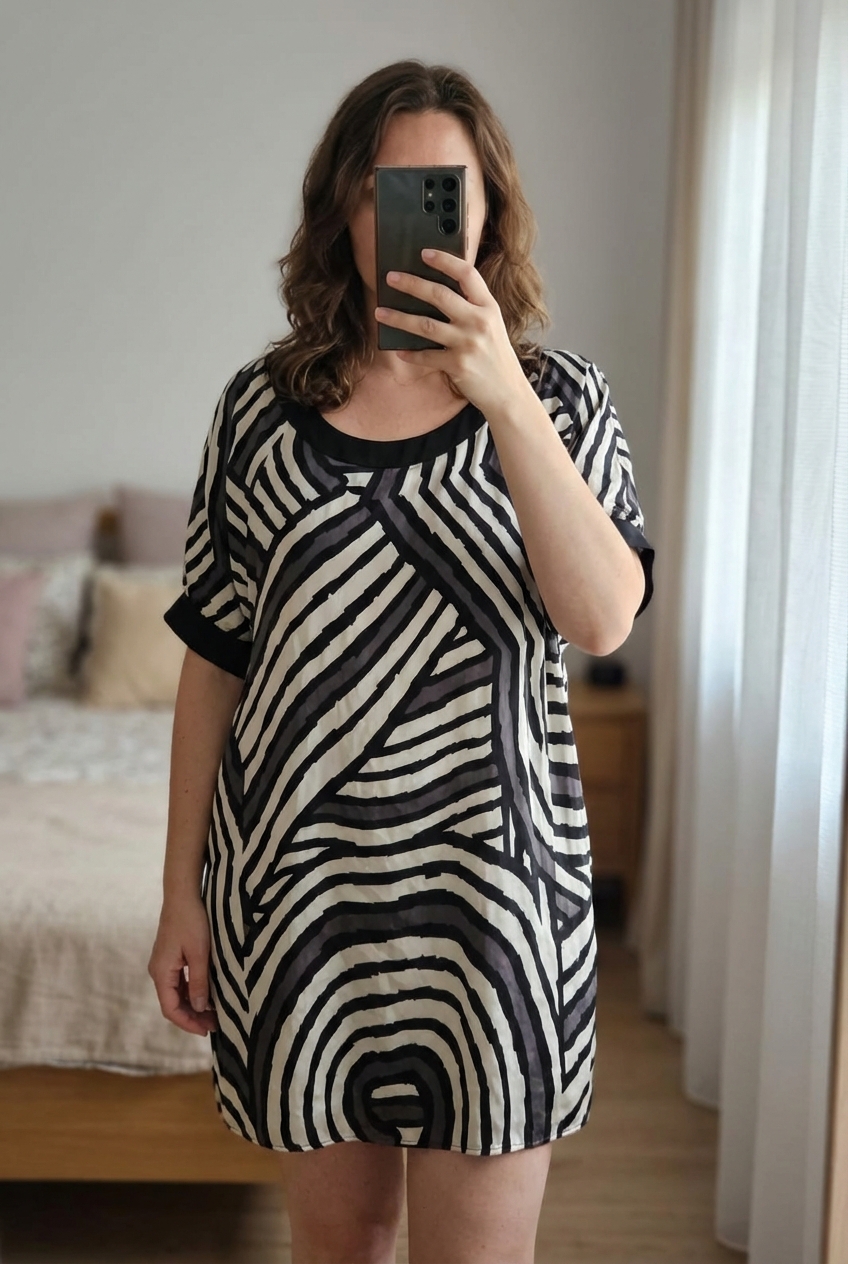 Zara robe tunique en satin imprimé gris blanc et noir debout, face à la caméra, dans une chambre à la lumière douce, avec une lumière naturelle filtrant par la fenêtre.