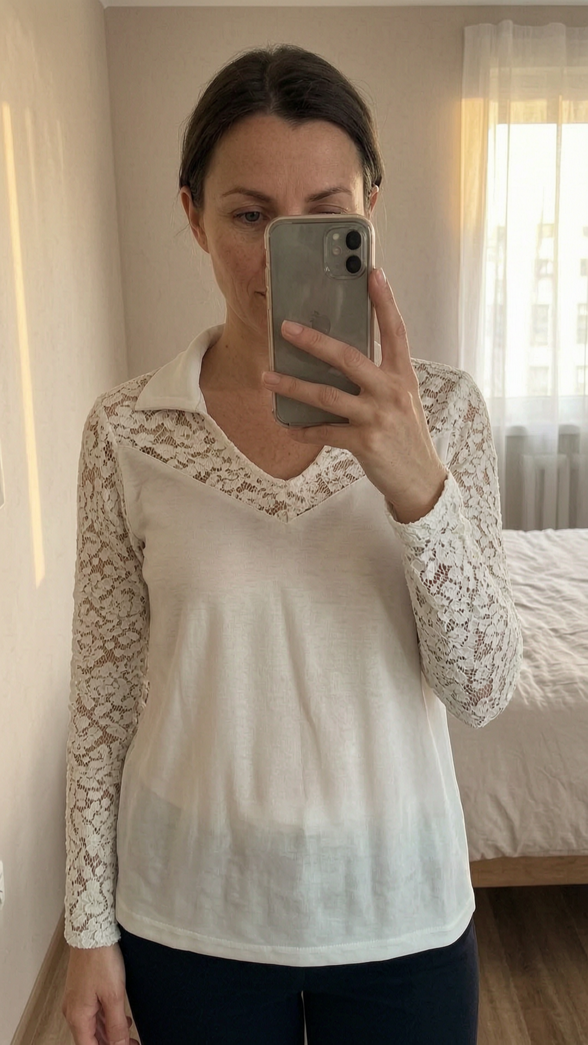 Blouse crème manches dentelle debout, face à la caméra, dans une chambre à la lumière douce, avec une lumière naturelle filtrant par la fenêtre.