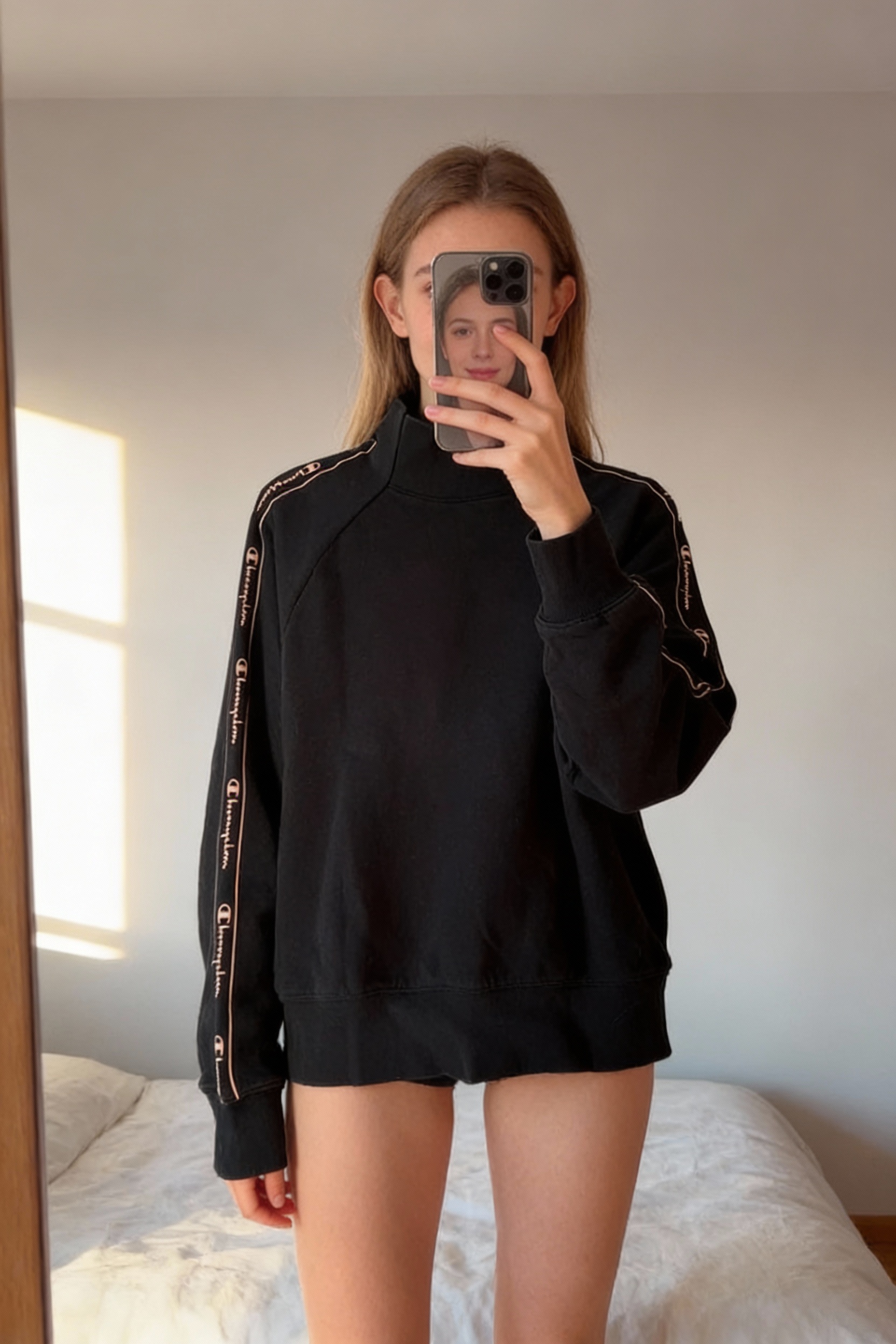 Una persona in Pull femme champion M - Maglione sport champion Donna in piedi, rivolto verso la fotocamera, in una camera da letto con luce soffusa e luce naturale che filtra dalla finestra.