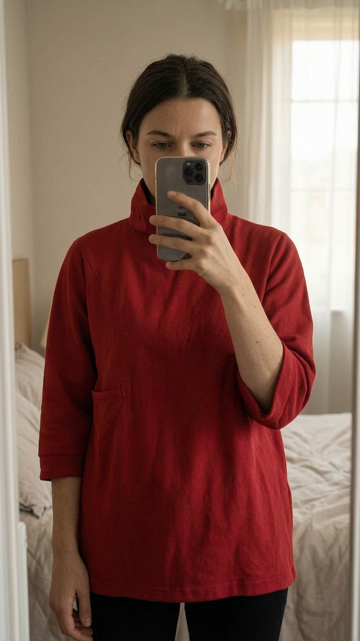 Haut rouge avec col et zip dans le dos debout, face à la caméra, dans une chambre à la lumière douce, avec une lumière naturelle filtrant par la fenêtre.