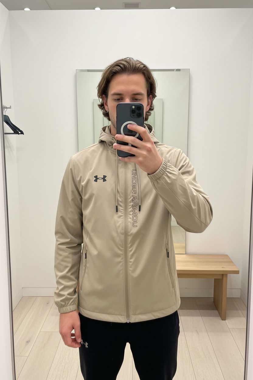 Une personne en Under Armour veste coupe-vent beige nouvelle collection debout, face à la caméra, dans une cabine d’essayage de style boutique avec un miroir et une décoration neutre.