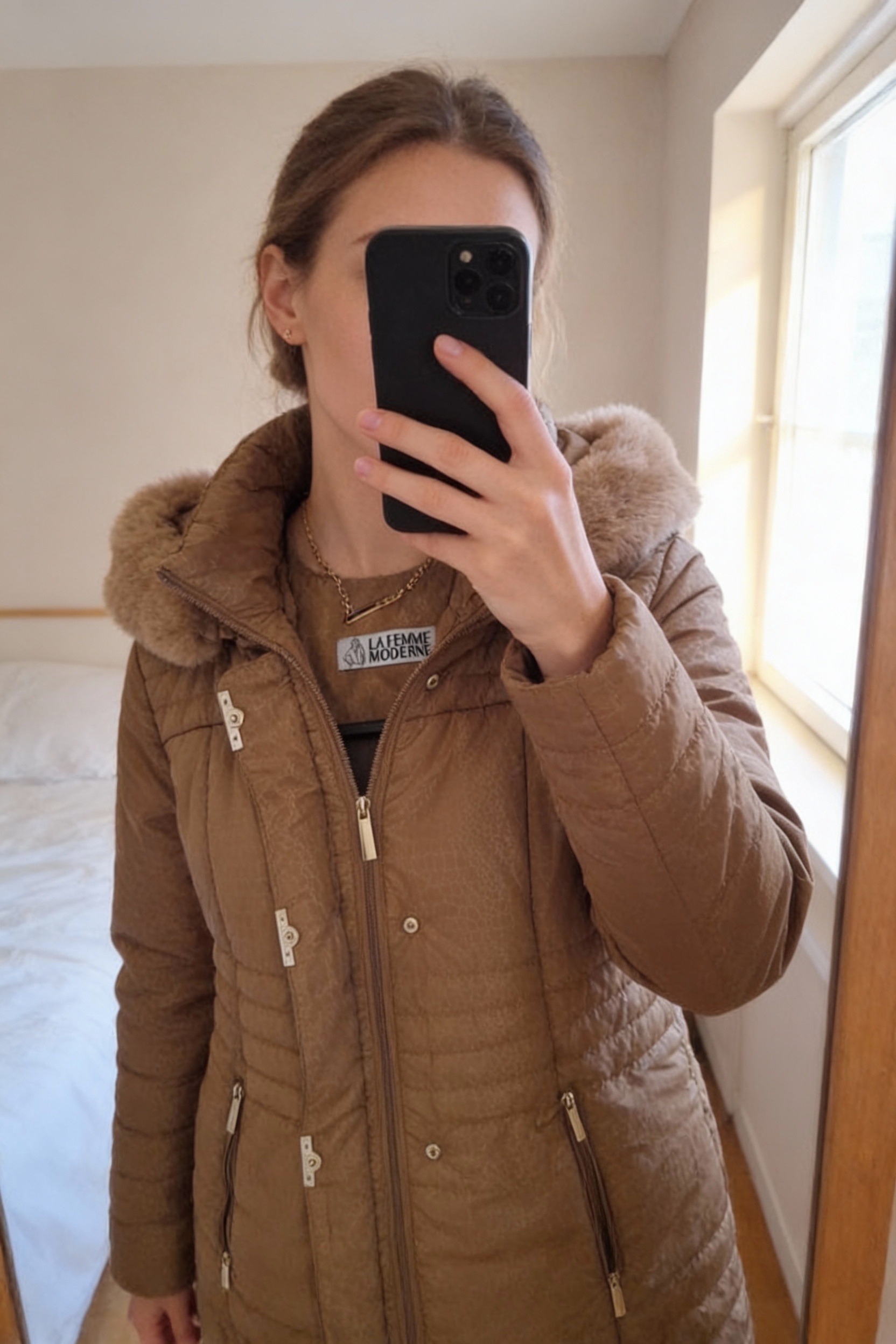 Manteau La femme moderne taille L debout, face à la caméra, dans une chambre à la lumière douce, avec une lumière naturelle filtrant par la fenêtre.