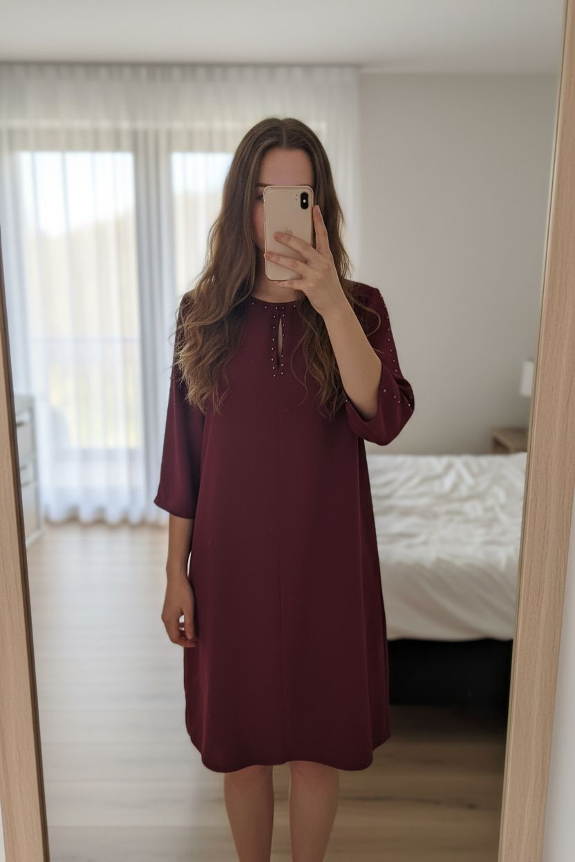 Robe légère bordeaux manches 3/4 taille 40 debout, face à la caméra, dans une chambre à la lumière douce, avec une lumière naturelle filtrant par la fenêtre.