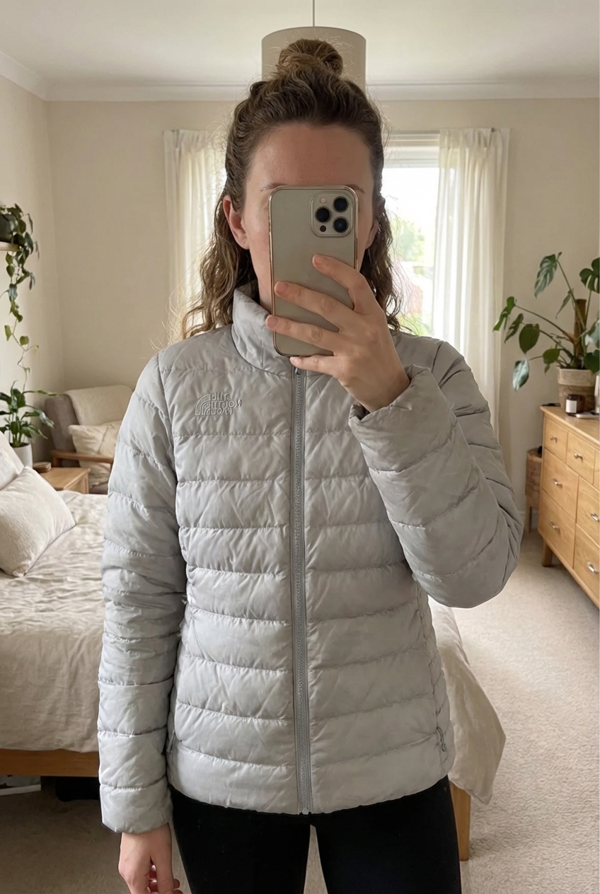Une personne en Doudoune Rembourré 550 HyVent Puffer Jacket The North Face Gris M debout, face à la caméra, dans une chambre à la lumière douce, avec une lumière naturelle filtrant par la fenêtre.