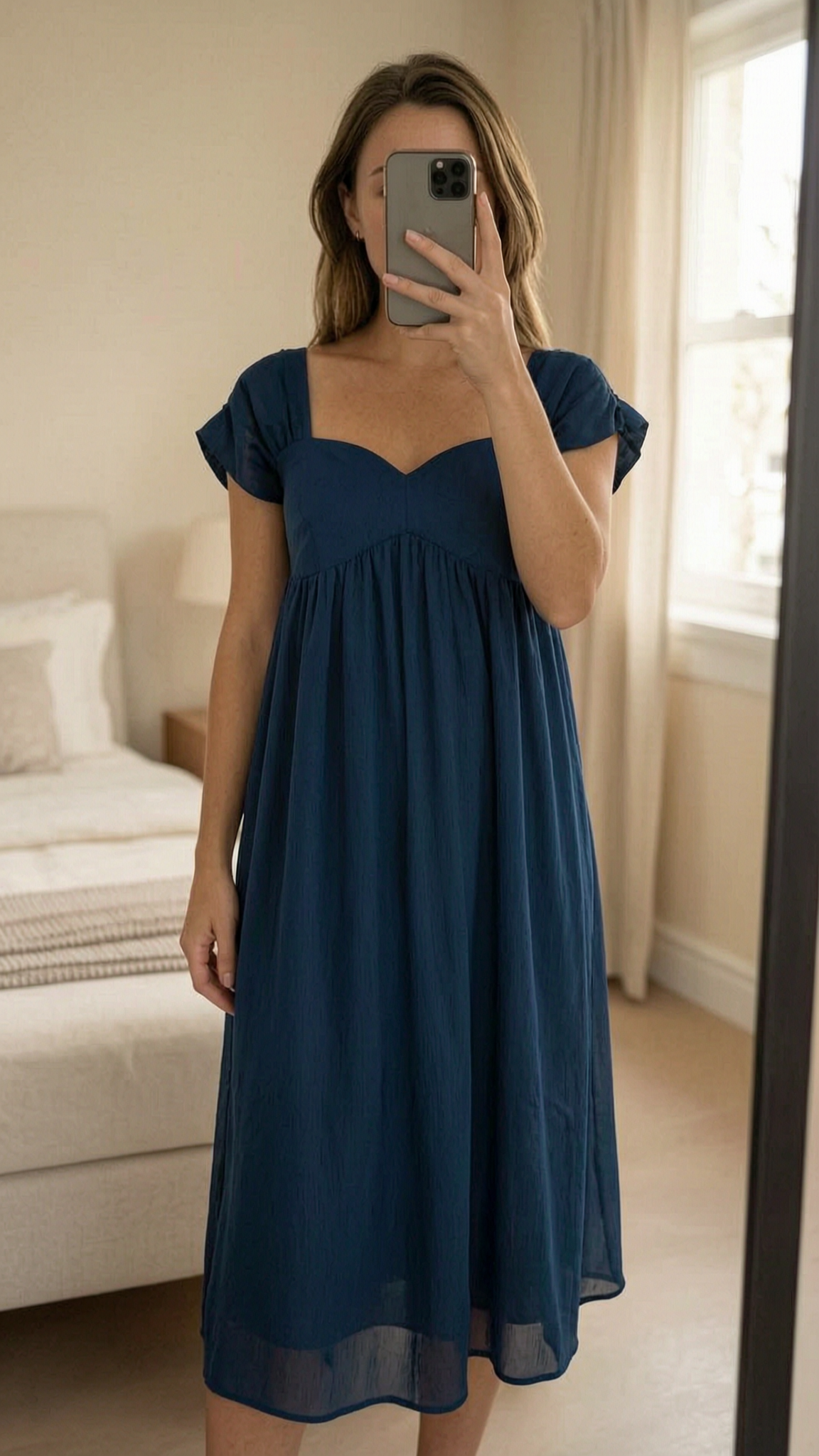 Robe trapèze Naf Naf debout, face à la caméra, dans une chambre à la lumière douce, avec une lumière naturelle filtrant par la fenêtre.