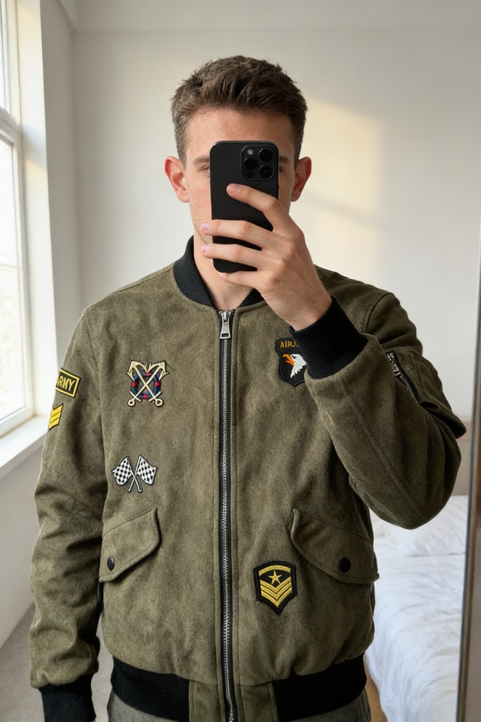 Veste Bombers en Daim Vintage Patch brodé Rembourré Kaki S debout, face à la caméra, dans une chambre à la lumière douce, avec une lumière naturelle filtrant par la fenêtre.