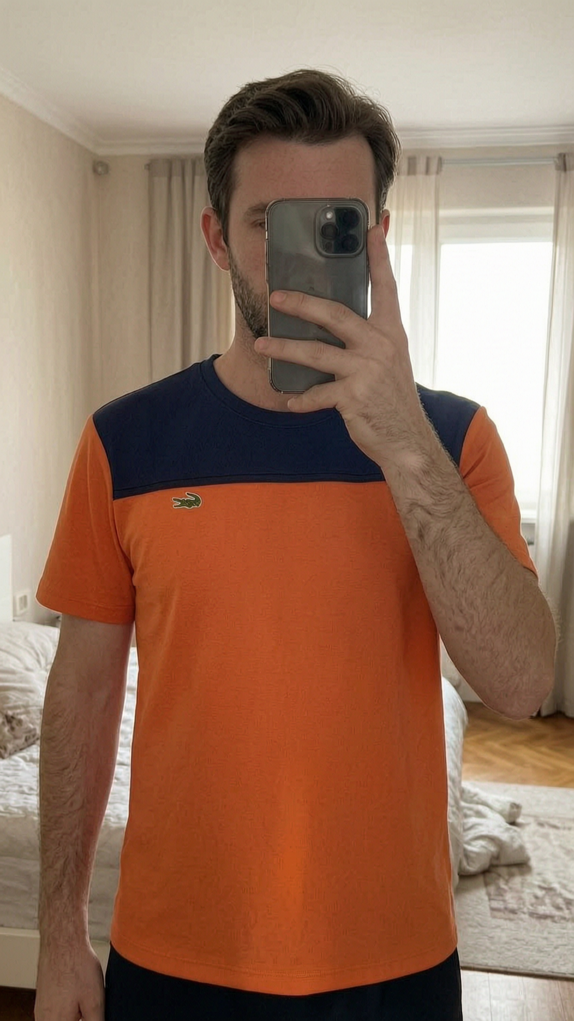 Tee-shirt Lacoste debout, face à la caméra, dans une chambre à la lumière douce, avec une lumière naturelle filtrant par la fenêtre.