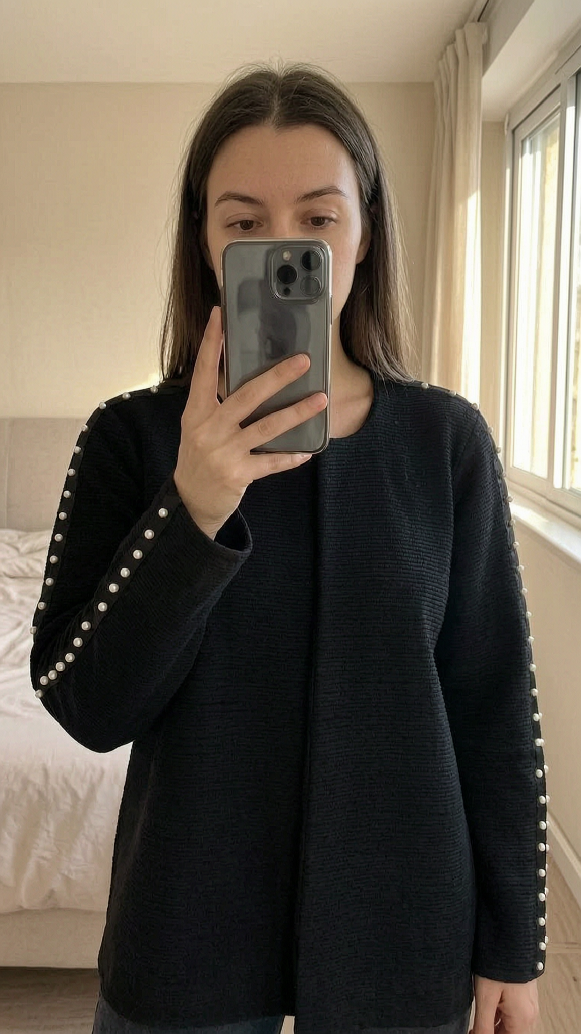 Veste sans bouton debout, face à la caméra, dans une chambre à la lumière douce, avec une lumière naturelle filtrant par la fenêtre.