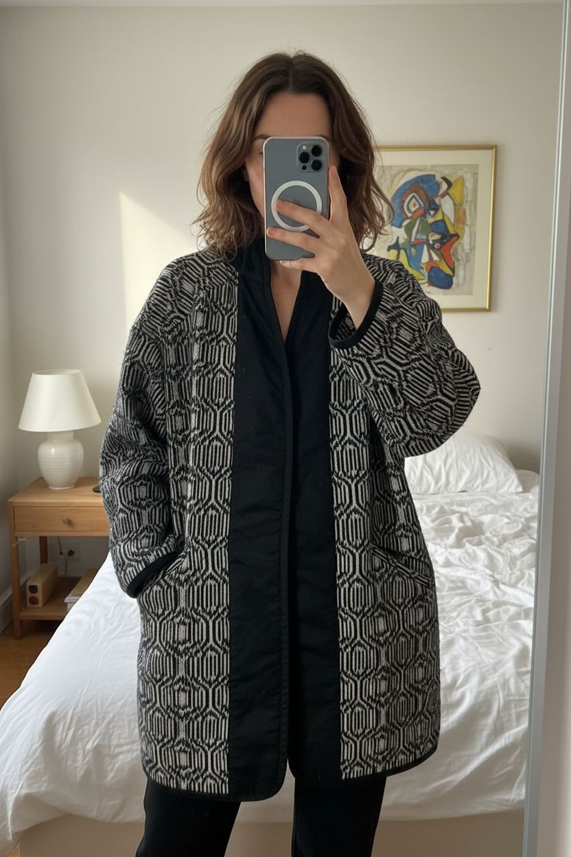 Veste longue mi saison debout, face à la caméra, dans une chambre à la lumière douce, avec une lumière naturelle filtrant par la fenêtre.