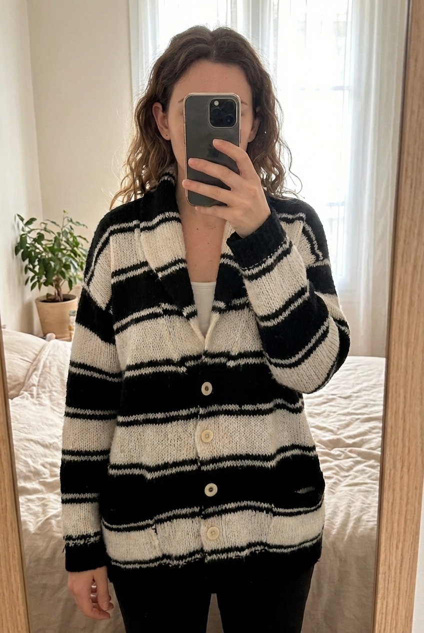 Gilet oversize debout, face à la caméra, dans une chambre à la lumière douce, avec une lumière naturelle filtrant par la fenêtre.