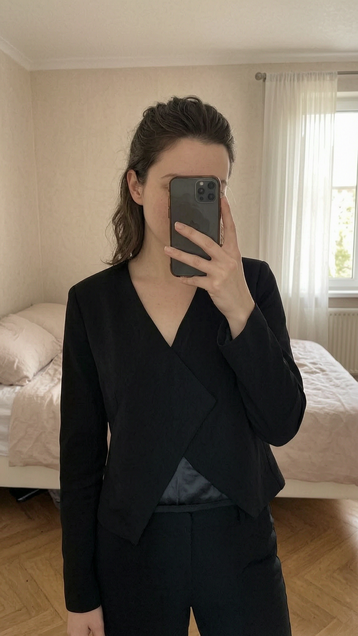 Petite veste noire mango debout, face à la caméra, dans une chambre à la lumière douce, avec une lumière naturelle filtrant par la fenêtre.