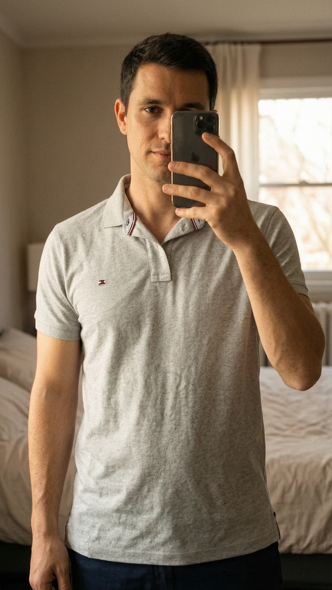 Tom Tailor Poloshirt hellgrau aufrecht stehend, der Kamera zugewandt, in einem sanft beleuchteten Schlafzimmer, mit natürlichem Licht, das durch ein Fenster fällt.