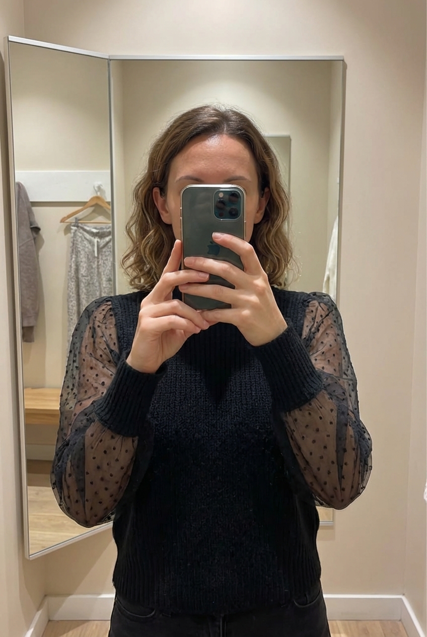 Une personne en Pull col roulé noir chic manches tulle pois transparents - État top ! debout, face à la caméra, dans une cabine d’essayage de style boutique avec un miroir et une décoration neutre.
