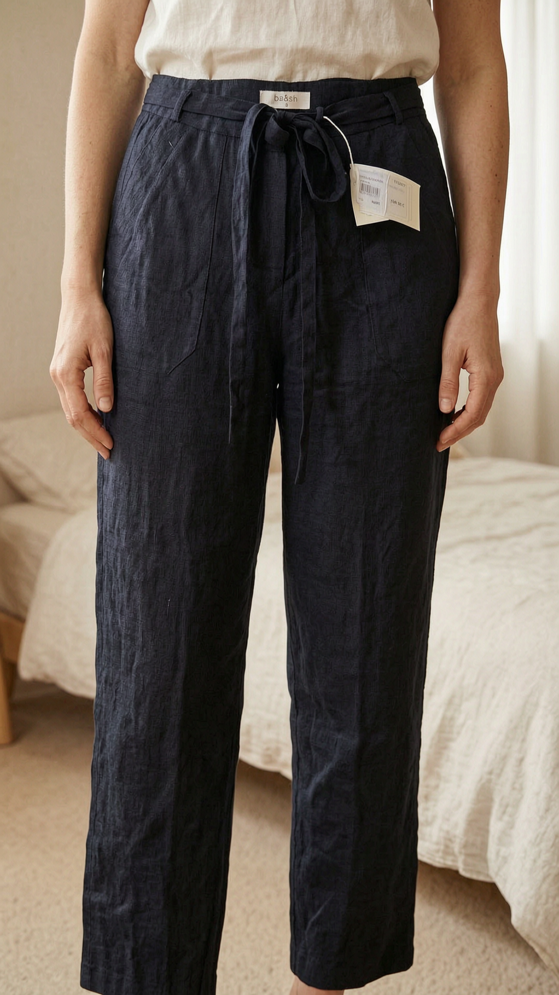 Bash linen style black pants with belt size 3 new w tags debout, face à la caméra, dans une chambre à la lumière douce, avec une lumière naturelle filtrant par la fenêtre.
