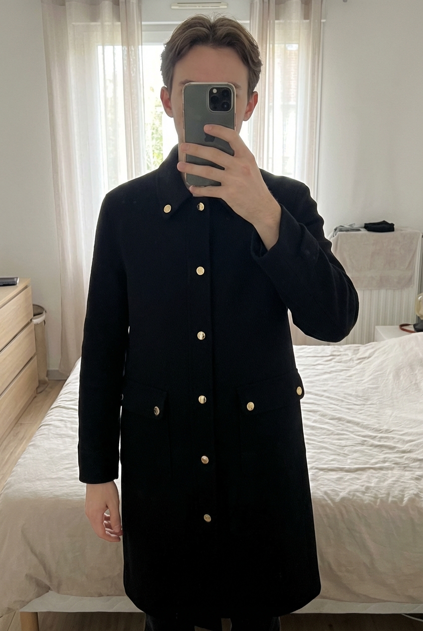 Une personne en Manteau Sandro debout, face à la caméra, avec une silhouette fine, dans une chambre à la lumière douce, avec une lumière naturelle filtrant par la fenêtre.