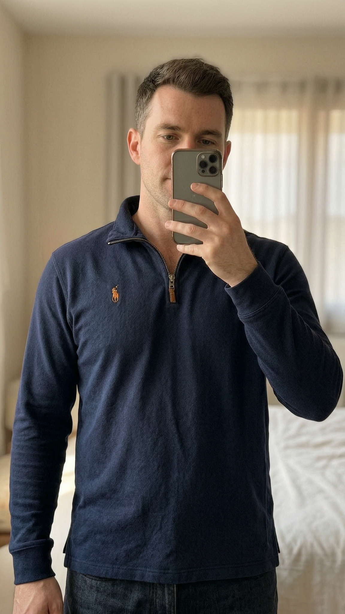 Polo homme debout, face à la caméra, dans une chambre à la lumière douce, avec une lumière naturelle filtrant par la fenêtre.
