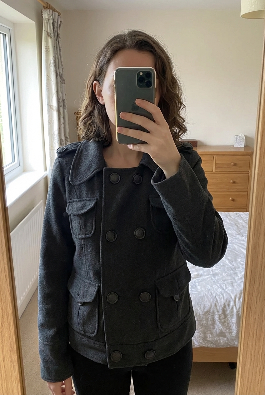 Veste courtes grise akoz debout, face à la caméra, dans une chambre à la lumière douce, avec une lumière naturelle filtrant par la fenêtre.