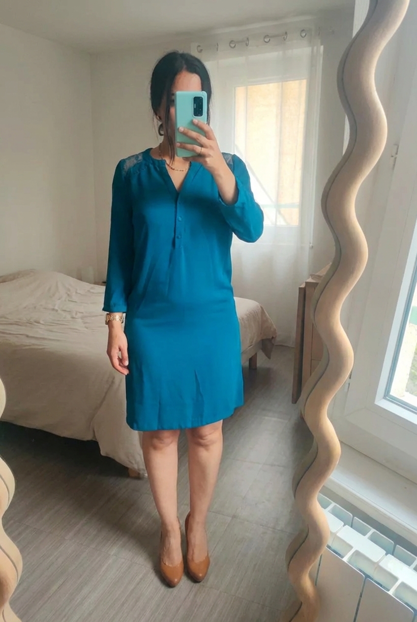 Robe bleu canard fluide et élégante dentelle debout, face à la caméra, dans une chambre à la lumière douce, avec une lumière naturelle filtrant par la fenêtre.