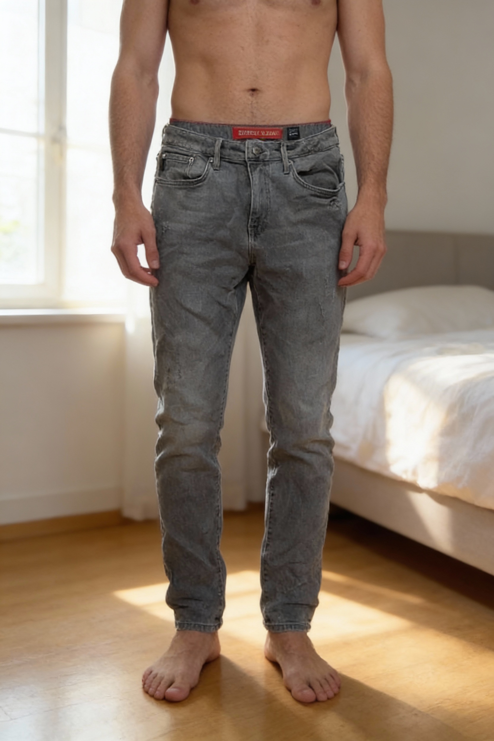 Jean superdry gris debout, face à la caméra, dans une chambre à la lumière douce, avec une lumière naturelle filtrant par la fenêtre.