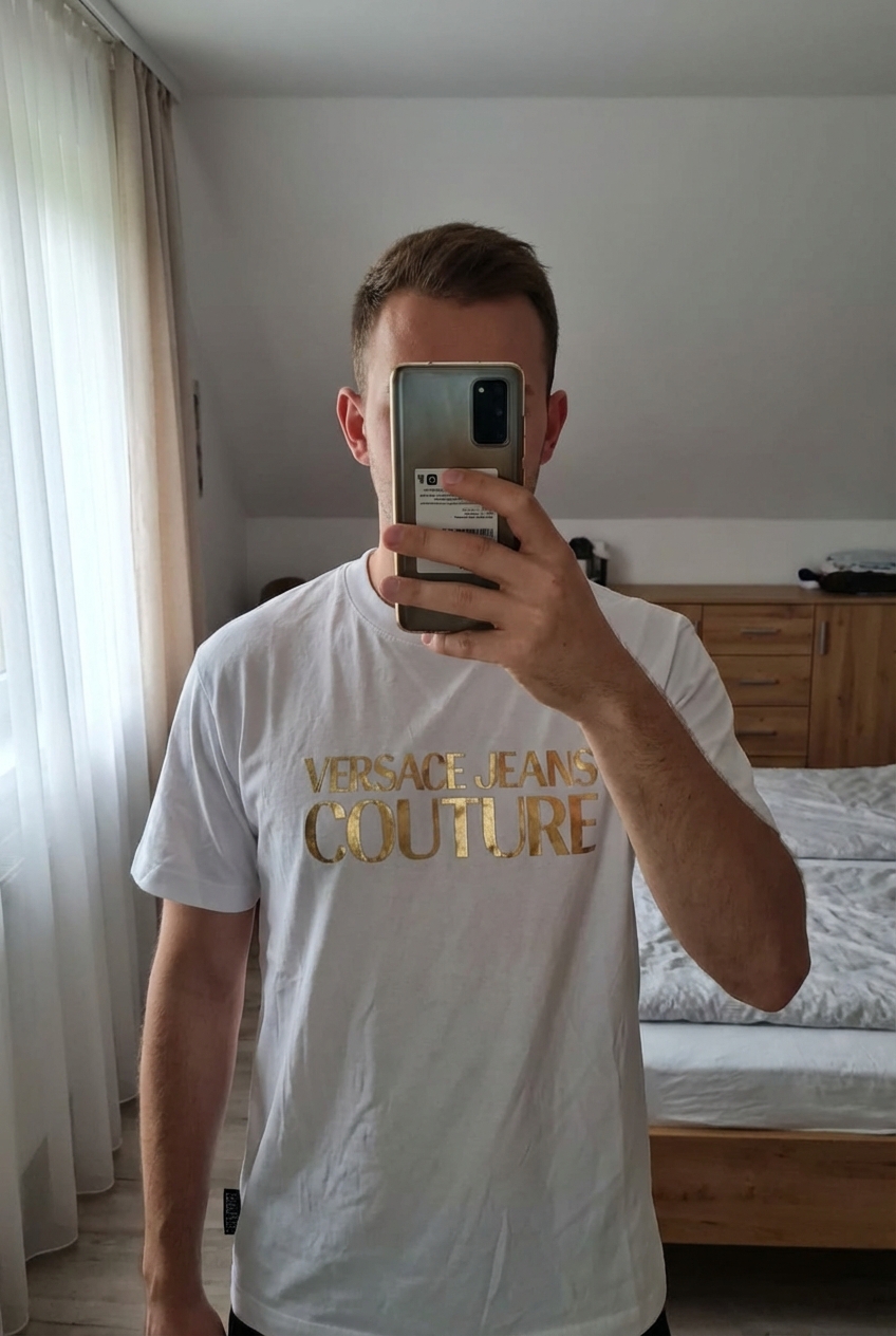 Tee-shirt versace debout, face à la caméra, dans une chambre à la lumière douce, avec une lumière naturelle filtrant par la fenêtre.