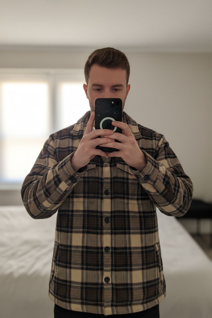 Veste style bucheron debout, face à la caméra, dans une chambre à la lumière douce, avec une lumière naturelle filtrant par la fenêtre.