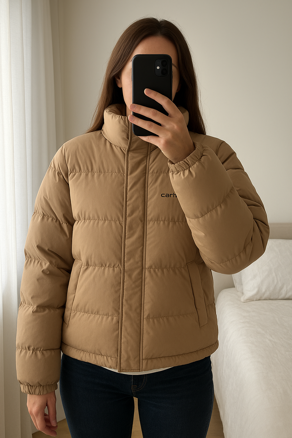 Doudoune Carharrt femme couleur sable debout, face à la caméra, dans une chambre à la lumière douce, avec une lumière naturelle filtrant par la fenêtre.