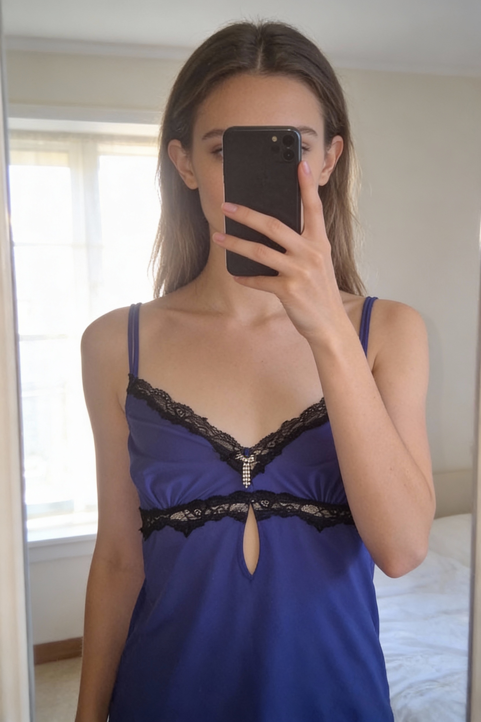 Caraco violet à dentelle noire debout, face à la caméra, dans une chambre à la lumière douce, avec une lumière naturelle filtrant par la fenêtre.