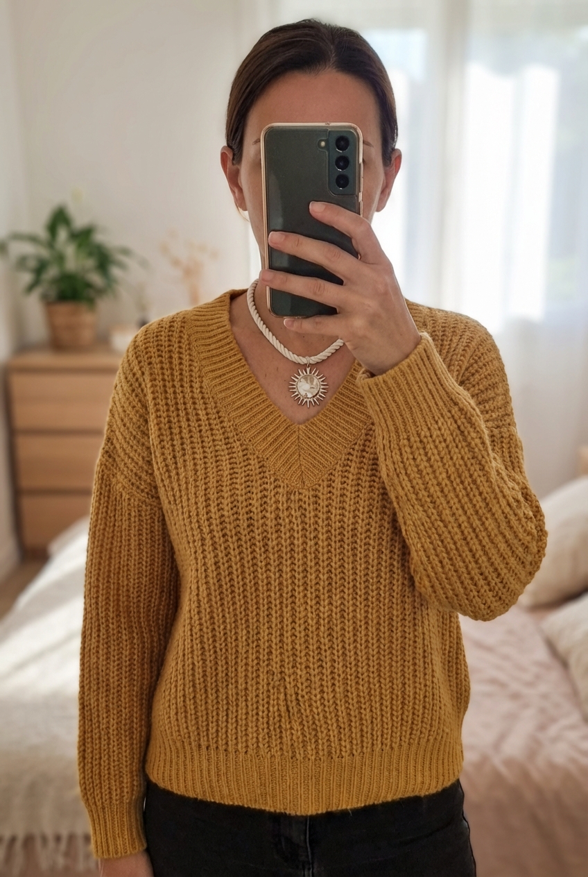 Une personne en Magnifique pull couleur moutarde – chaud et élégant pour l’automne/hiver debout, face à la caméra, dans une chambre à la lumière douce, avec une lumière naturelle filtrant par la fenêtre.