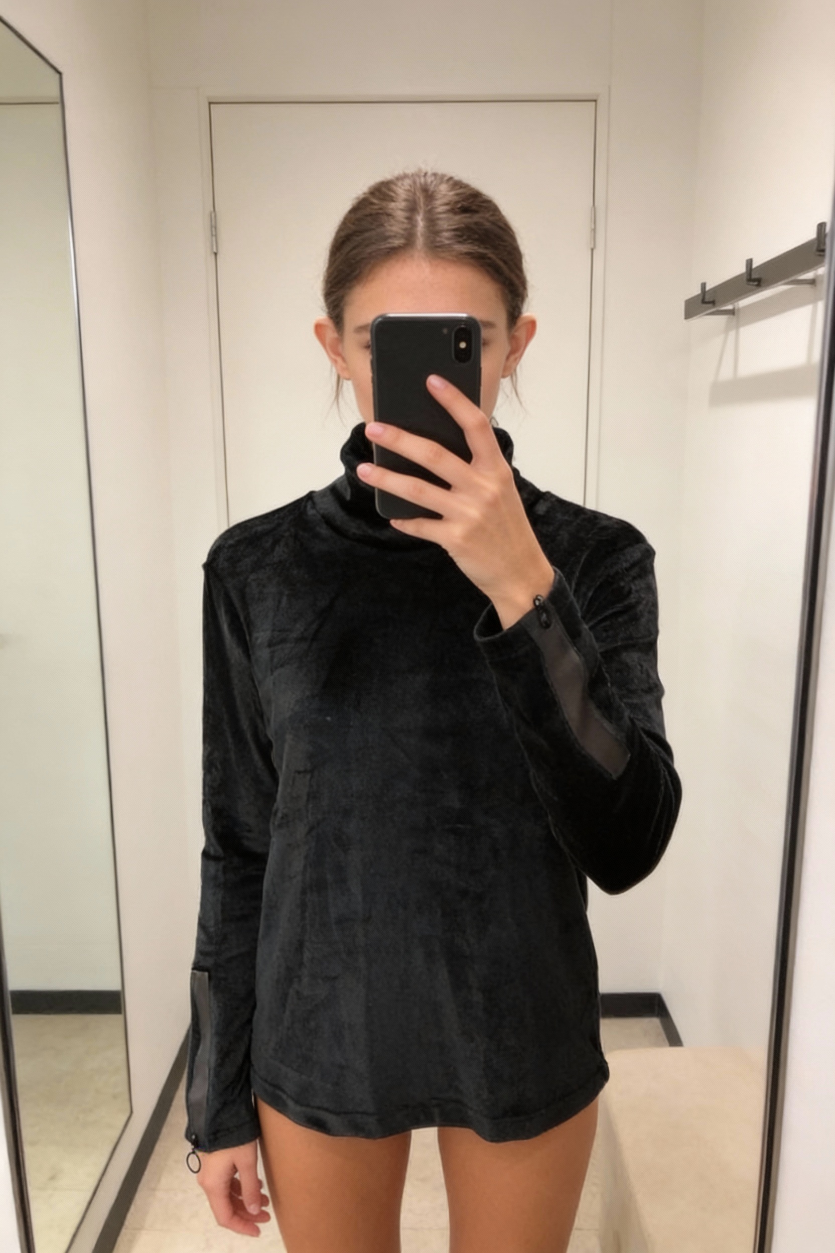 Une personne en Haut en velours darkwear debout, face à la caméra, dans une cabine d’essayage de style boutique avec un miroir et une décoration neutre.