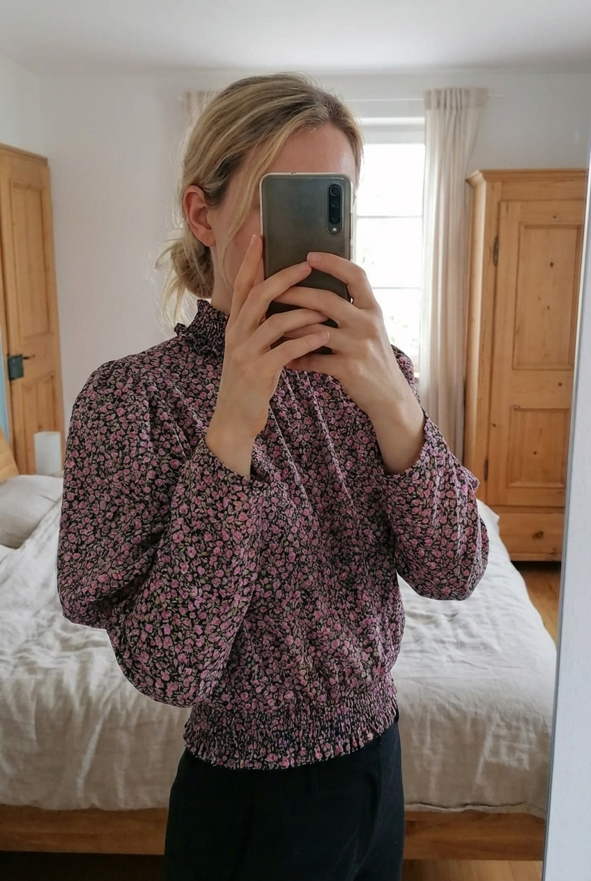 Chemisier à fleurs Zara TBE debout, face à la caméra, dans une chambre à la lumière douce, avec une lumière naturelle filtrant par la fenêtre.