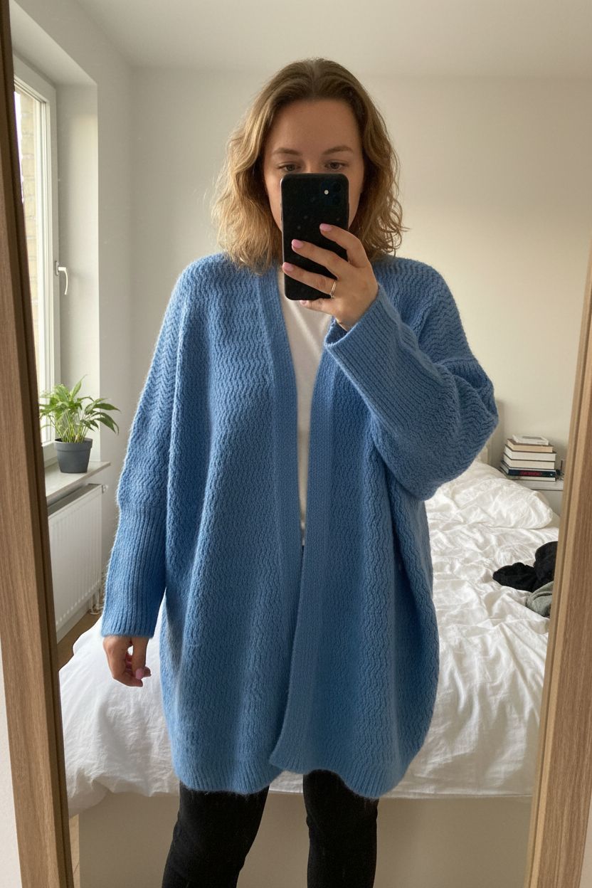 Eine Person in more & more strickjacke oversized aufrecht stehend, der Kamera zugewandt, mit etwas kurvigeren Körperformen, in einem sanft beleuchteten Schlafzimmer, mit natürlichem Licht, das durch ein Fenster fällt.