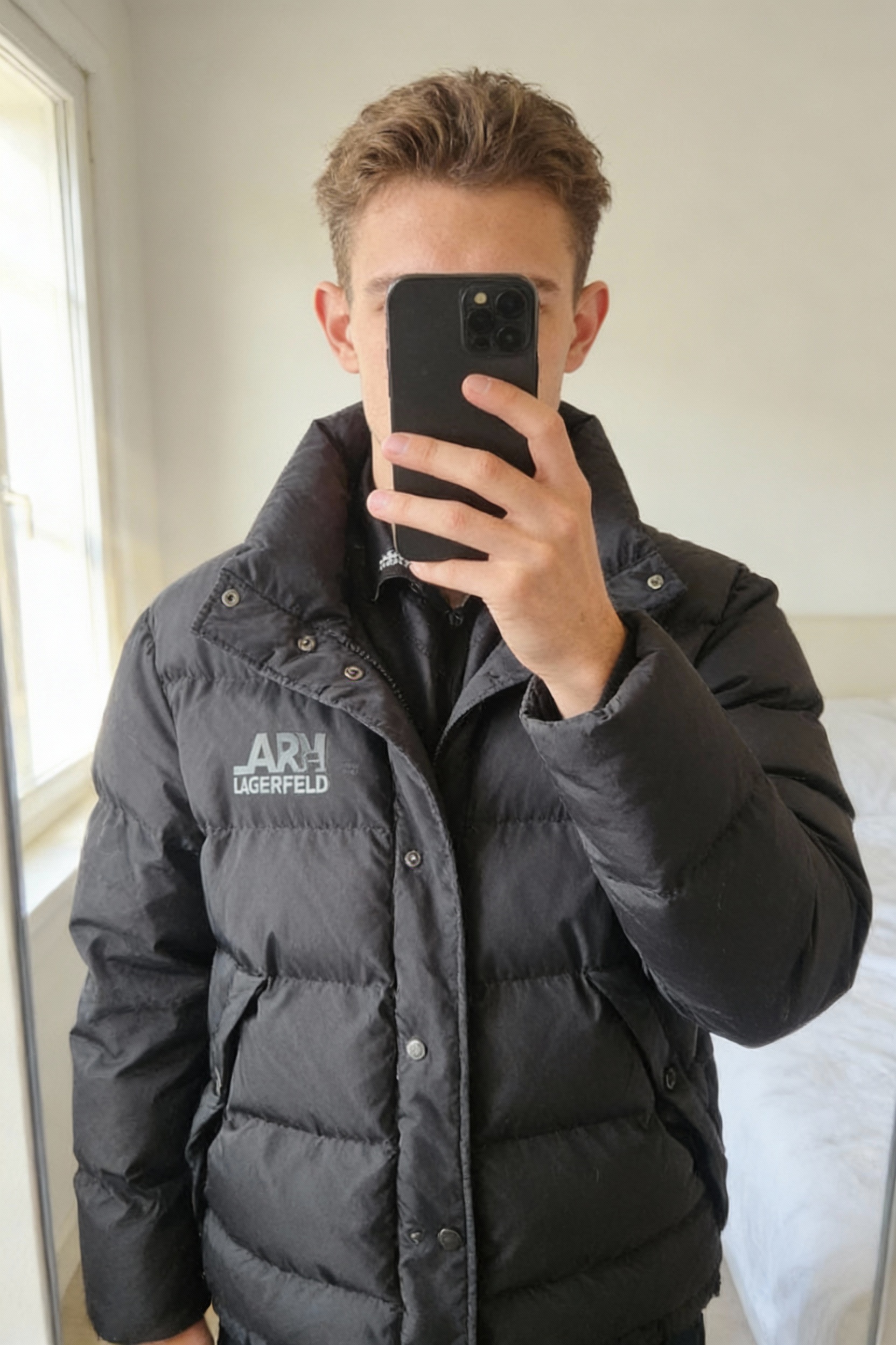 Doudoune Karl Lagerfeld noir debout, face à la caméra, dans une chambre à la lumière douce, avec une lumière naturelle filtrant par la fenêtre.