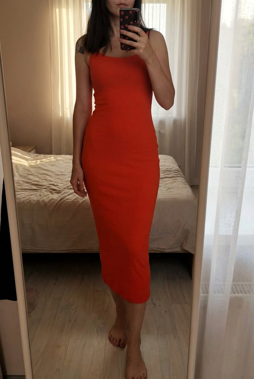 Robe longue moulante H&M orange sanguine Taille XS 🧡 debout, face à la caméra, dans une chambre à la lumière douce, avec une lumière naturelle filtrant par la fenêtre.
