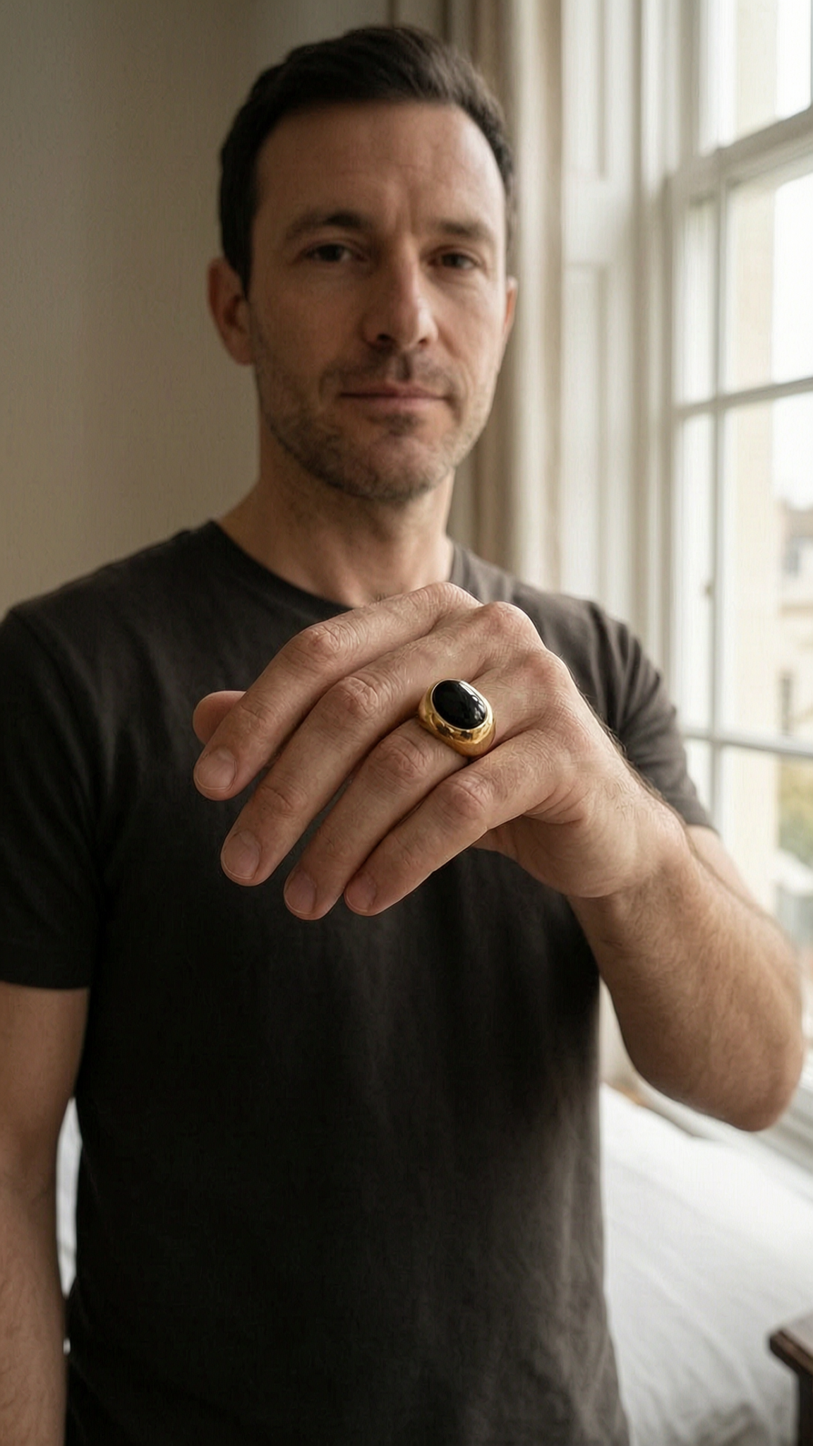 Chevalière onyx plaqué or debout, face à la caméra, dans une chambre à la lumière douce, avec une lumière naturelle filtrant par la fenêtre.