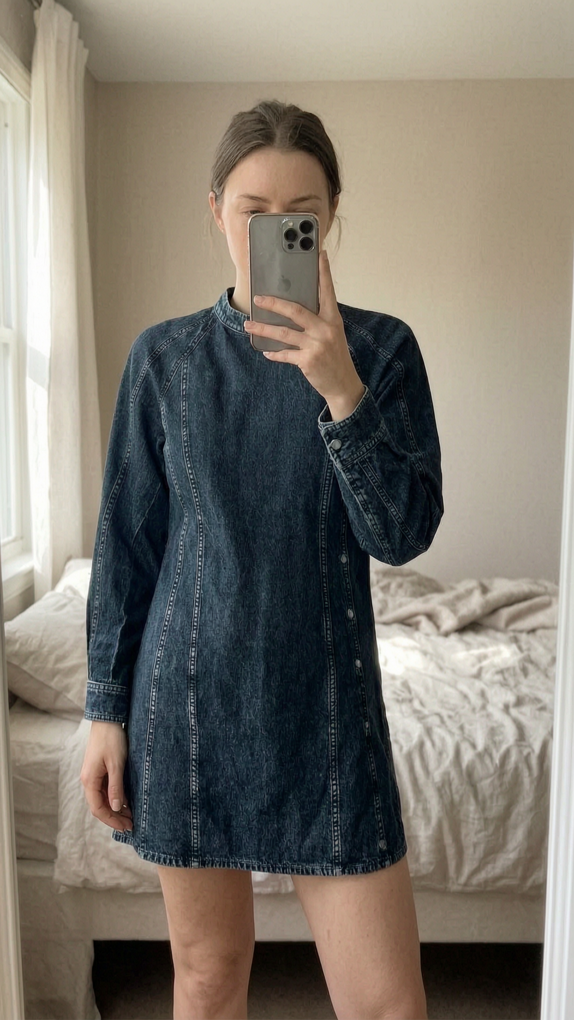 & Other Stories Black Denim Dress standing straight facing the camera, in a softly lit bedroom setting, with gentle natural light filtering through a window.