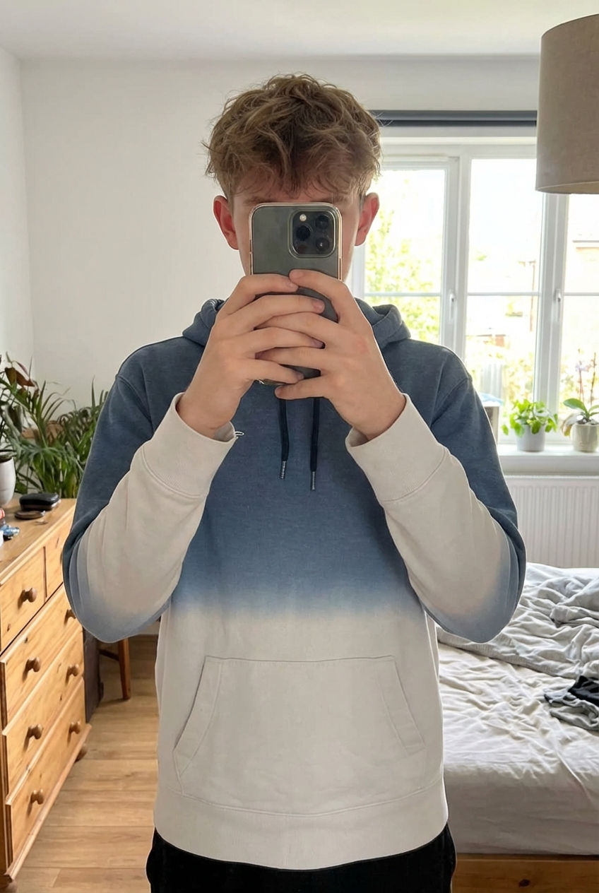 Sweat bleu et blanc à capuche Hollister debout, face à la caméra, dans une chambre à la lumière douce, avec une lumière naturelle filtrant par la fenêtre.