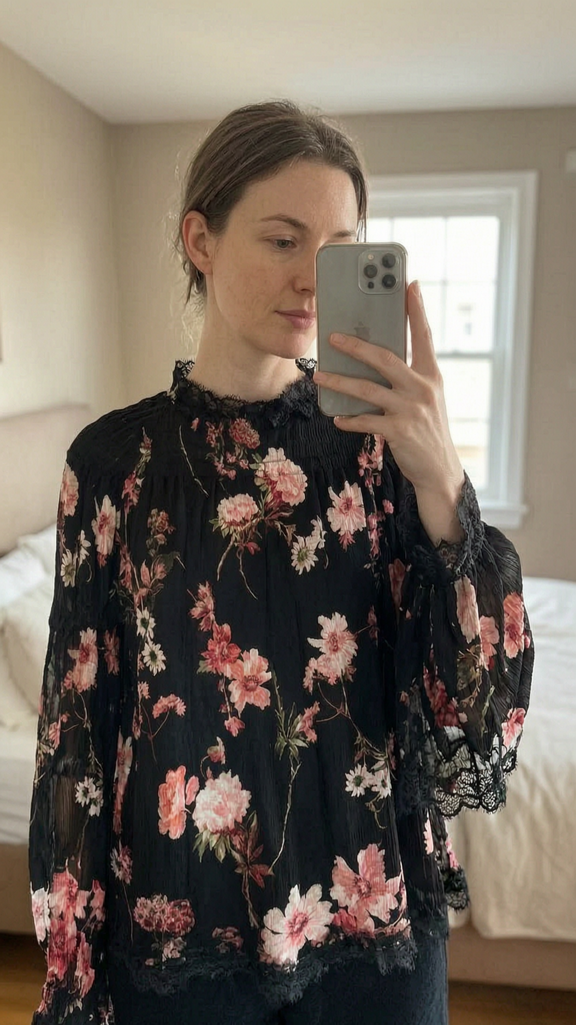 Haut The Kooples taille 1 soie debout, face à la caméra, dans une chambre à la lumière douce, avec une lumière naturelle filtrant par la fenêtre.