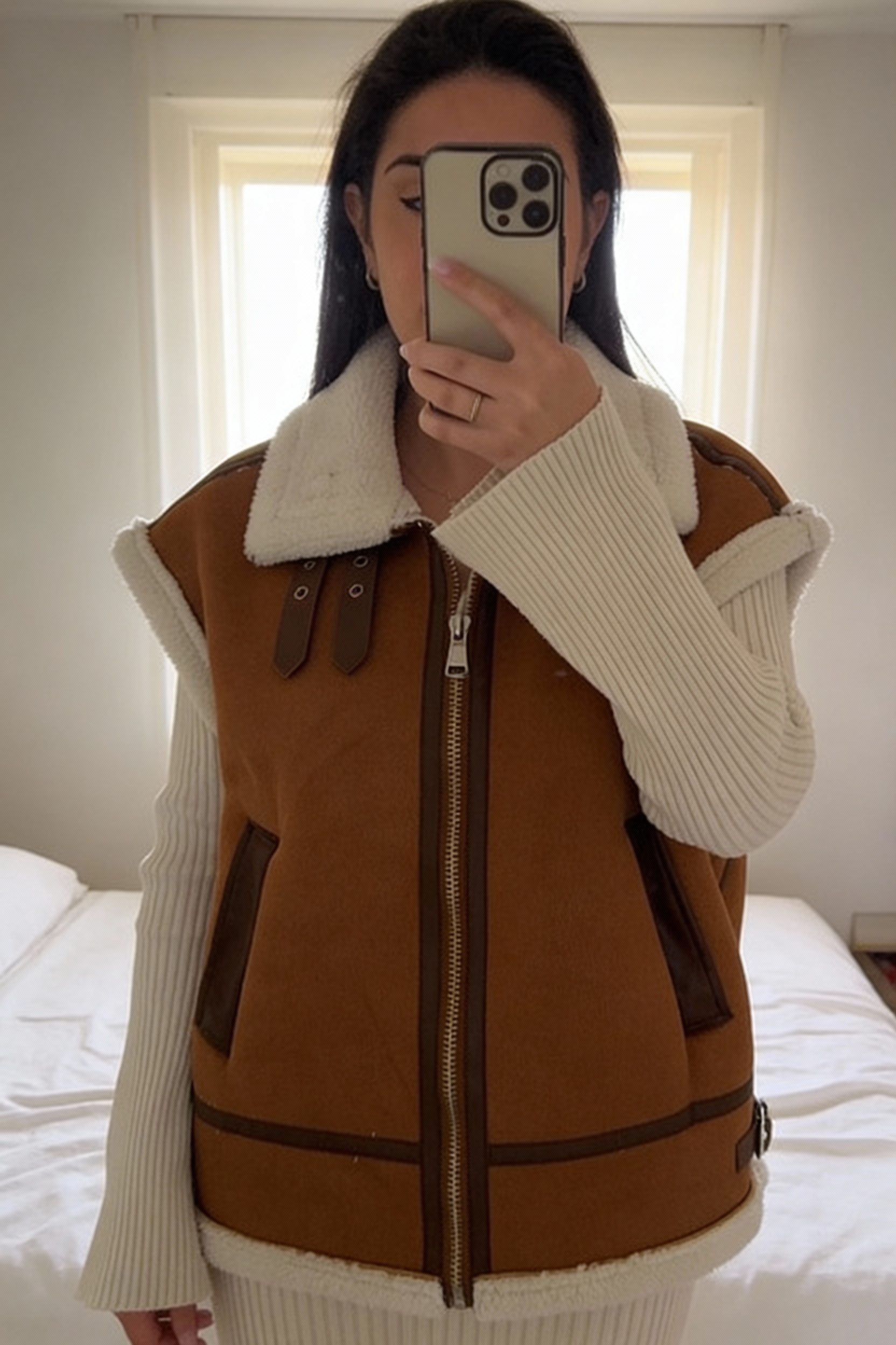 Manteau sans manches à fourrure debout, face à la caméra, dans une chambre à la lumière douce, avec une lumière naturelle filtrant par la fenêtre.