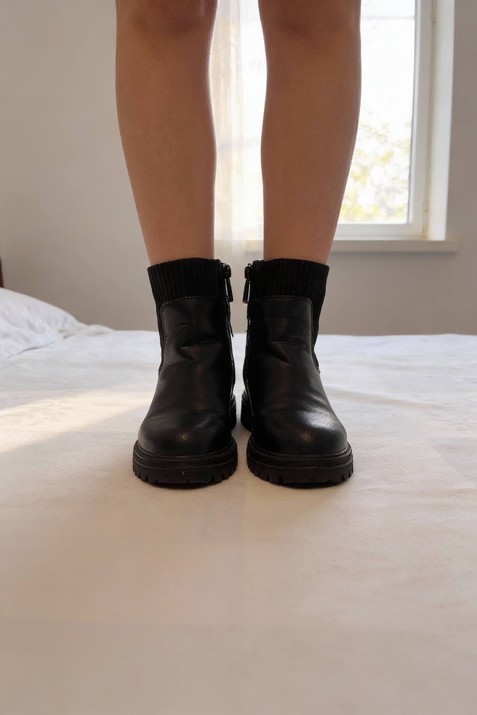 Boots Fille unies a col Chaussette debout, face à la caméra, dans une chambre à la lumière douce, avec une lumière naturelle filtrant par la fenêtre.