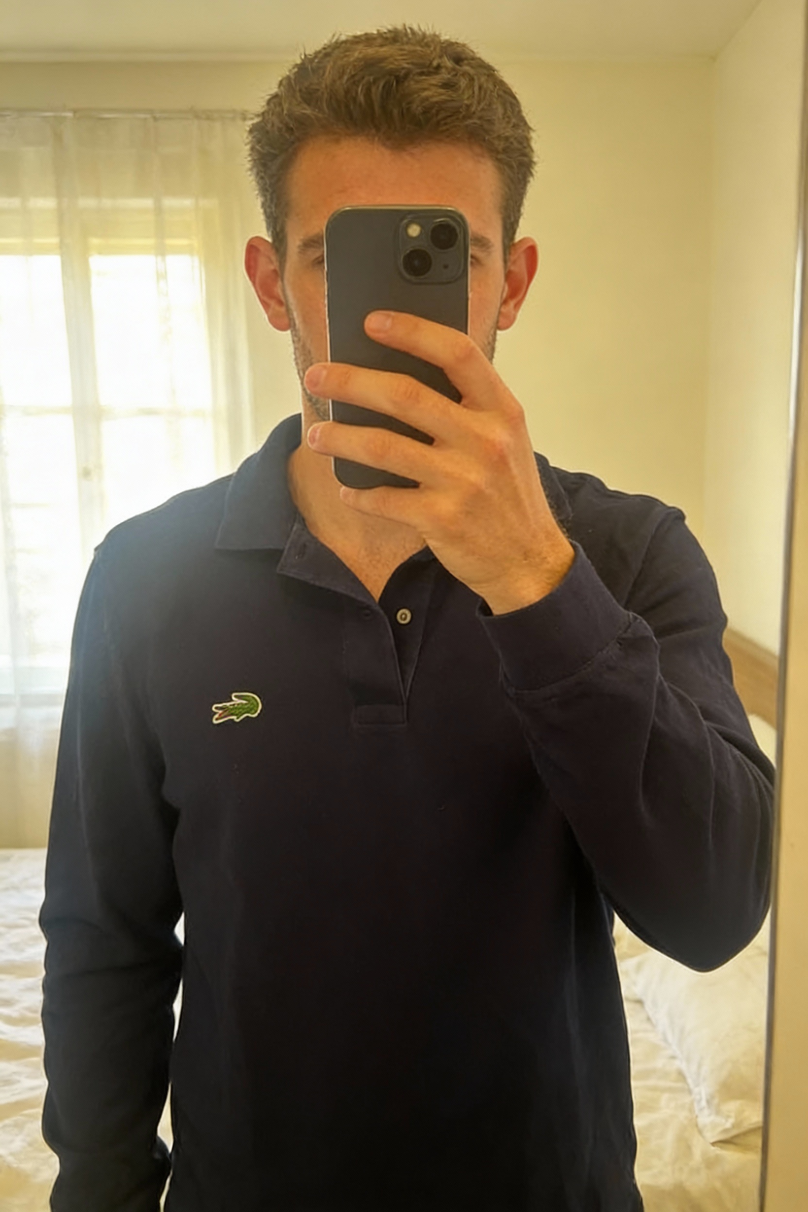 Lacoste long sleeve polo shirt navy blue - Size S men debout, face à la caméra, dans une chambre à la lumière douce, avec une lumière naturelle filtrant par la fenêtre.