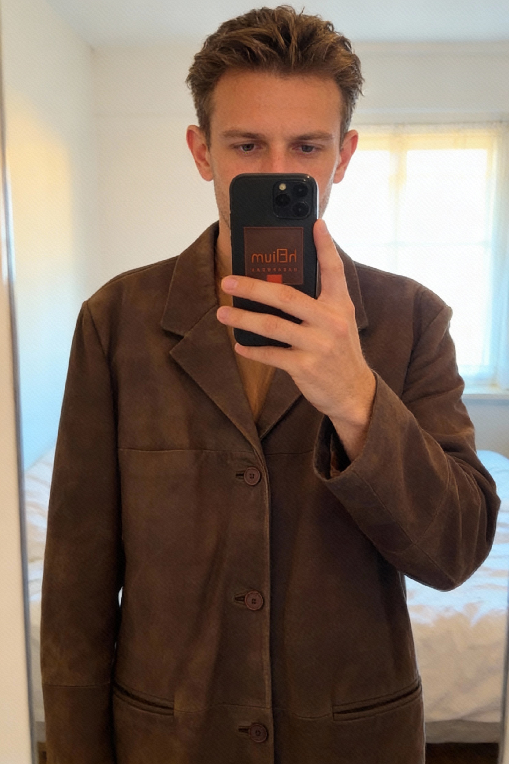 Veste Homme en Cuir Nubuck Marron debout, face à la caméra, dans une chambre à la lumière douce, avec une lumière naturelle filtrant par la fenêtre.